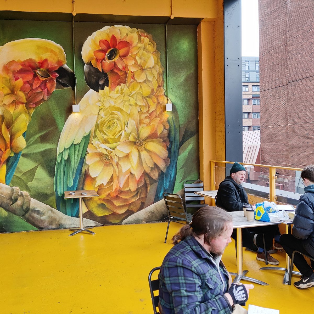 sketchers sat on a yellow balcony in front of a giant mural of exotic birds made of flowers.
